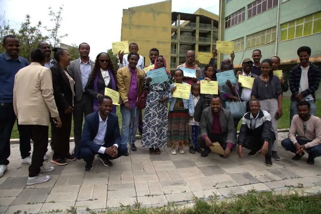 A group portrait outdoors featuring people holding up Certificate of Completion papers in front of a multi-story building.