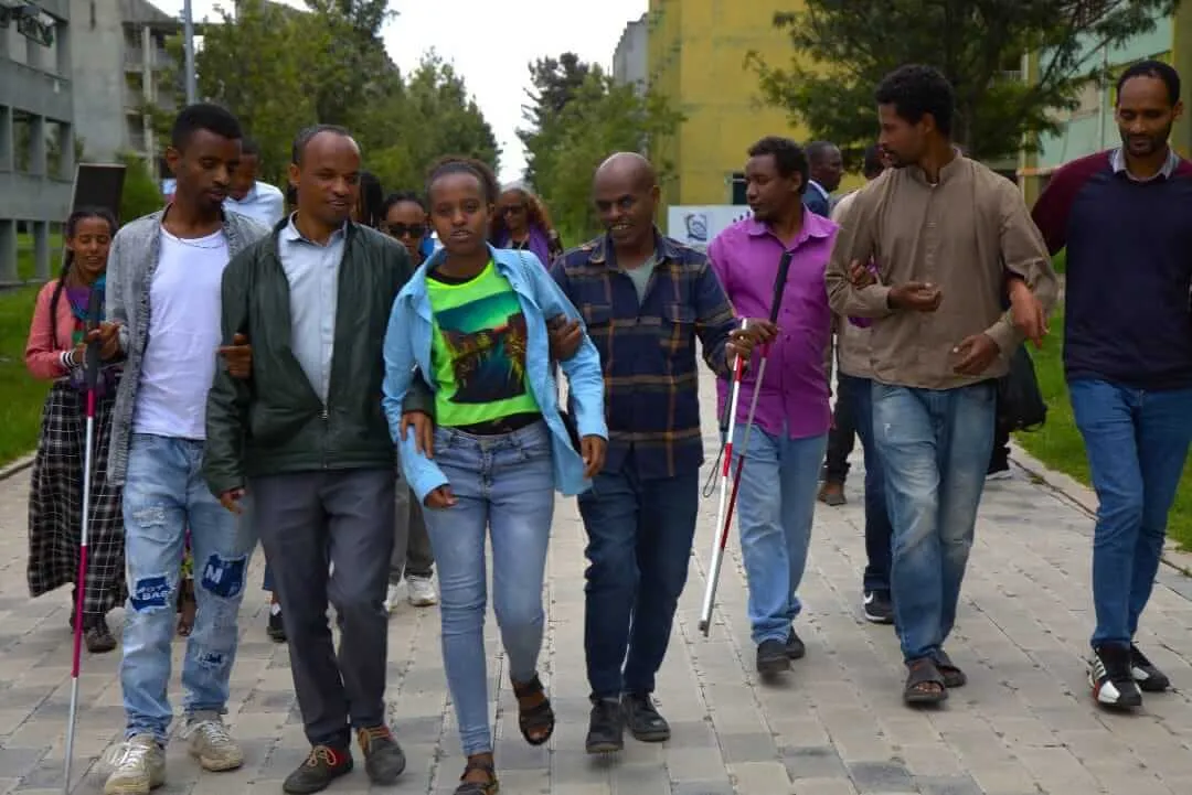 A group of people walking together along a paved outdoor walkway, with individuals holding white canes for guidance.