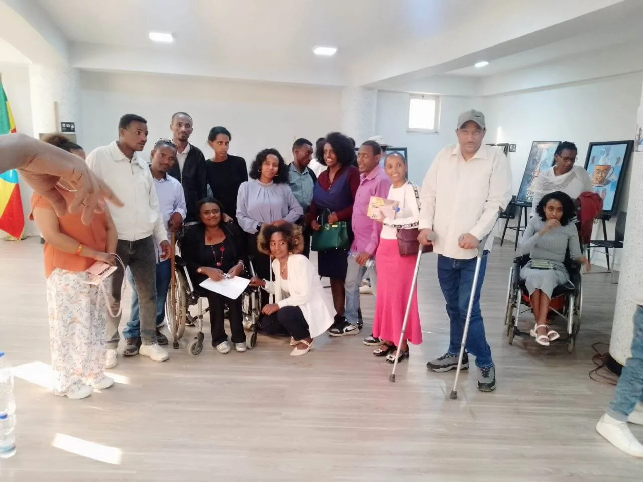 A indoor group gathering with participants standing and seated, including wheelchair users and individuals with mobility aids, posing for a group picture in an exhibition space.