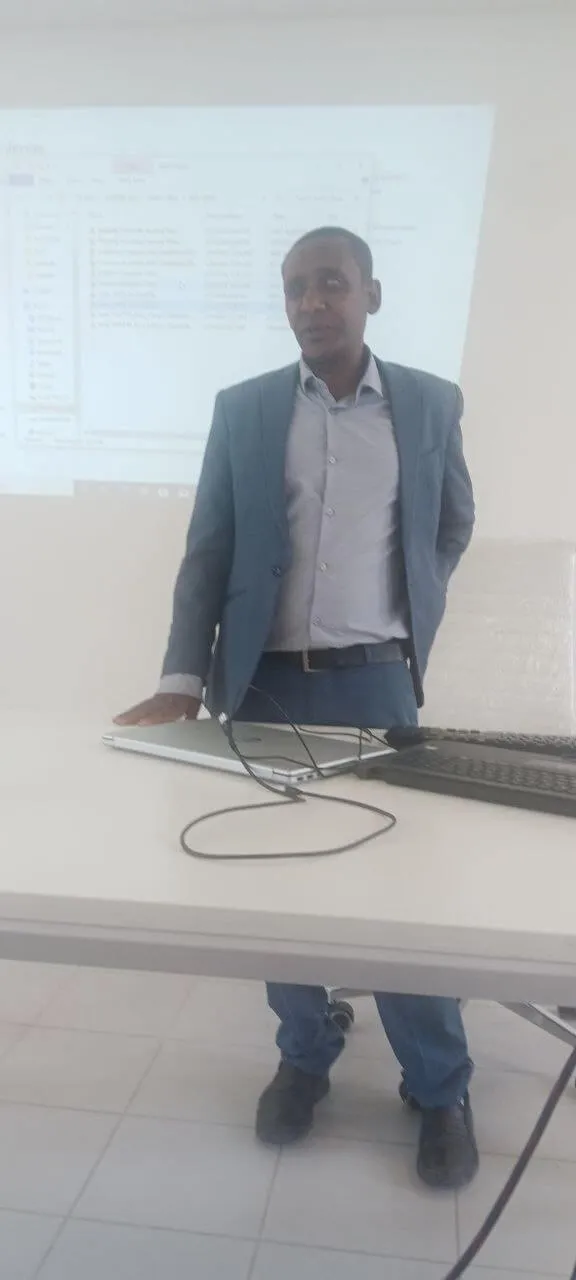A presenter wearing a blue suit jacket standing near a table with laptops in front of a projection screen displaying a digital folder menu.