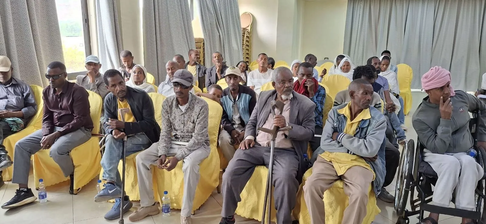 An audience seated in yellow-covered chairs inside a hall, listening attentively during an event or presentation.